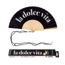 Ventaglio pieghevole nero con stecche in legno chiaro. Sul tessuto nero è stampata in bianco la frase 'la dolce vita'. Un cordino nero è appoggiato sotto il ventaglio, accanto alla sua scatola di imballaggio coordinata che riporta la stessa frase e il logo 'fisura'."