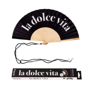 Ventaglio pieghevole nero con stecche in legno chiaro. Sul tessuto nero è stampata in bianco la frase 'la dolce vita'. Un cordino nero è appoggiato sotto il ventaglio, accanto alla sua scatola di imballaggio coordinata che riporta la stessa frase e il logo 'fisura'."
