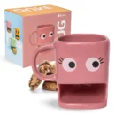 "Tazza in ceramica rosa di forma squadrata con due grandi occhi bianchi, lunghe ciglia nere e una fessura rettangolare per i biscotti alla base. Dietro la tazza ci sono alcuni biscotti con gocce di cioccolato e, sulla sinistra, la scatola di cartone della tazza con la scritta 'fisura. MUG COOKIE' e un'immagine di tazze di vari colori."