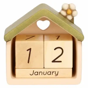 Calendario da tavolo THUN a forma di casetta in ceramica color crema con tetto verde e apertura a cuore, contenente cubi di legno con numeri e mese "January", decorato con un fiore bianco sul tetto.