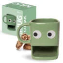Tazza in ceramica verde chiaro di forma squadrata con due grandi occhi bianchi e neri sul davanti e una fessura rettangolare per i biscotti alla base. Dietro la tazza ci sono alcuni biscotti con gocce di cioccolato e, sulla sinistra, la scatola di cartone verde della tazza con la scritta 'fisura. MUG COOKIE