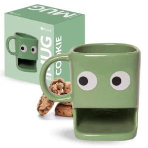 Tazza in ceramica verde chiaro di forma squadrata con due grandi occhi bianchi e neri sul davanti e una fessura rettangolare per i biscotti alla base. Dietro la tazza ci sono alcuni biscotti con gocce di cioccolato e, sulla sinistra, la scatola di cartone verde della tazza con la scritta 'fisura. MUG COOKIE