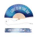 Ventaglio pieghevole con tessuto a gradiente che sfuma dal blu scuro all'azzurro chiaro, con stecche in legno chiaro. Sul tessuto è stampata in bianco la frase 'LOVE IS IN THE AIR'. Un cordino bianco è adagiato sotto il ventaglio, accanto alla sua scatola di imballaggio coordinata che riporta la stessa frase e il logo 'fisura'