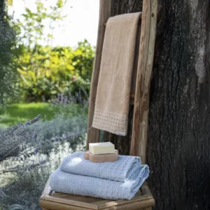 Set 2 asciugamani Iconic Elegance THUN azzurro in cotone 100%, morbidi e raffinati, piegati su un tavolino accanto a una scala in legno con asciugamano beige.
